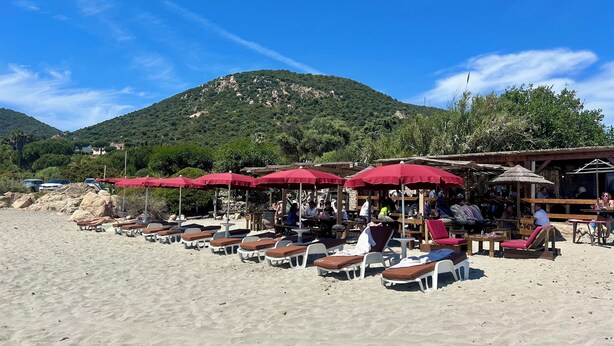 A beachside area with sun loungers and red umbrellas, a wooden structure with tables and chairs, and a mountainous backdrop.