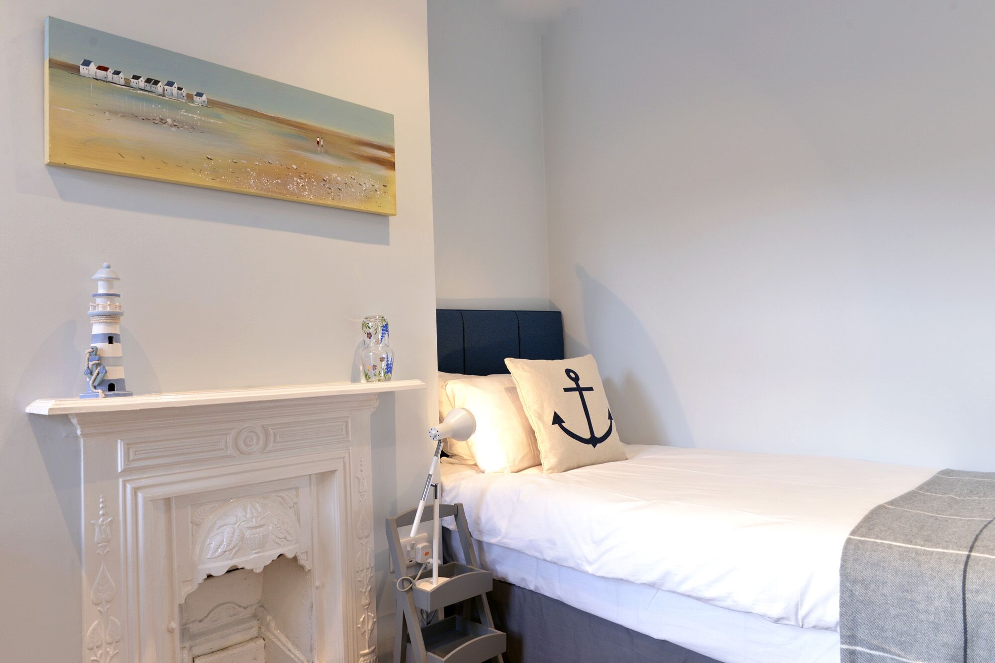 A bedroom with a bed, a nightstand, a lamp, a painting, a fireplace, and a decorative lighthouse.