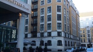 Um edifício de vários andares com uma placa indicando "DOUBLETREE BY HILTON" e "NATURAL KITCHEN" no andar térreo. Possui áreas de estar ao ar livre com mesas e cadeiras, e vários carros estacionados em frente.