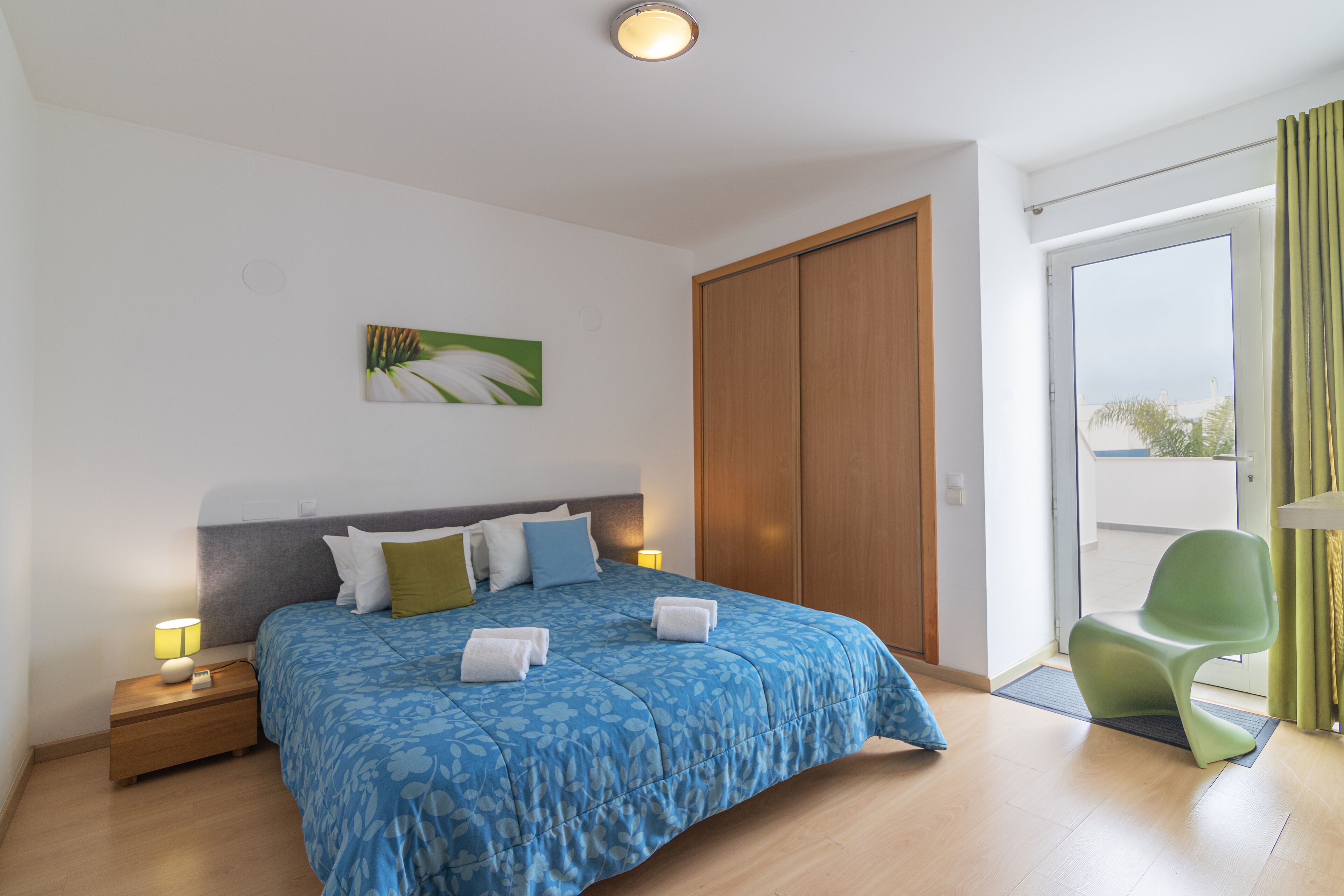 A modern hotel room with a large bed, a green chair, and a view through a sliding door.