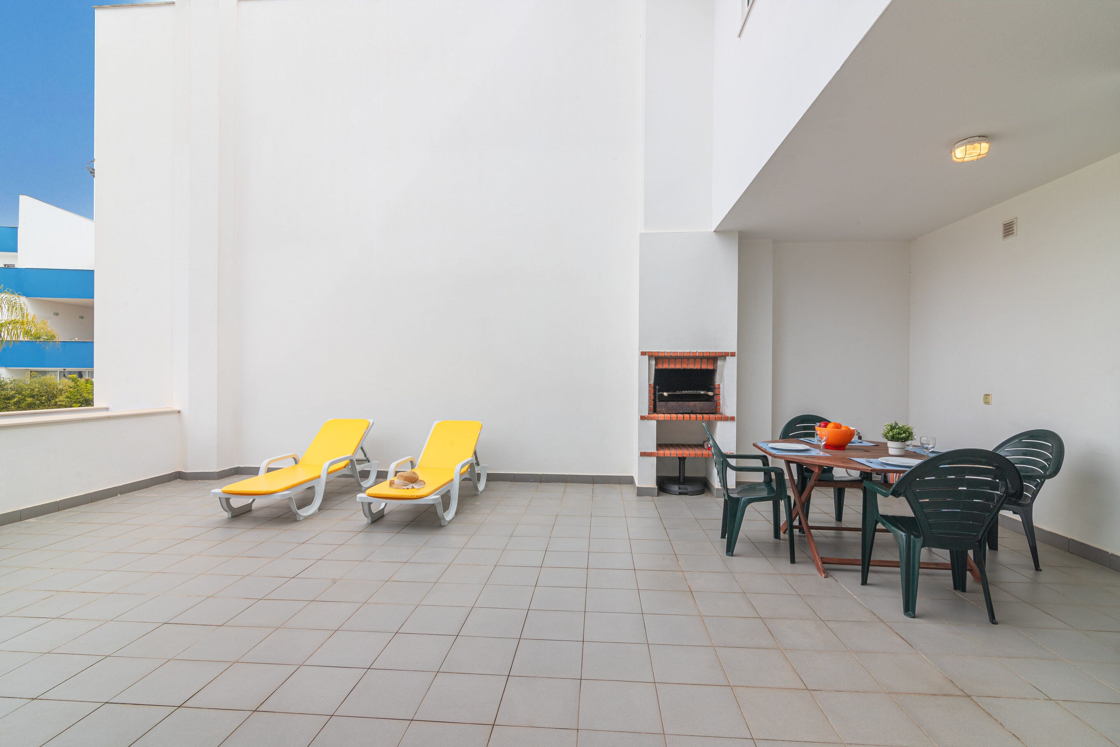 Une terrasse extérieure ensoleillée, avec deux chaises longues jaunes, une table avec des chaises et un barbecue.