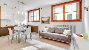 Una cocina moderna con armarios blancos, una mesa de comedor con sillas, un sofá gris y ventanas amplias con marcos de madera.