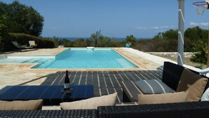 A pool area with lounge chairs and a table, a basketball hoop, and a view of the ocean. - Stylish luxury trulli with private pool and amazing sea view  (Ostuni)