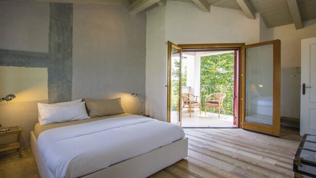 A bedroom with a large bed, a nightstand, a lamp, and a sliding glass door leading to an outdoor seating area with chairs. - Capriva Del Friuli: House / Villa - Capriva del Friuli (Capriva del Friuli)