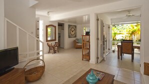 A spacious living area with a television, wooden furniture, and a view of a tropical garden through an open door. - Charming townhouse only 10 minutes from the beach! (Porters)
