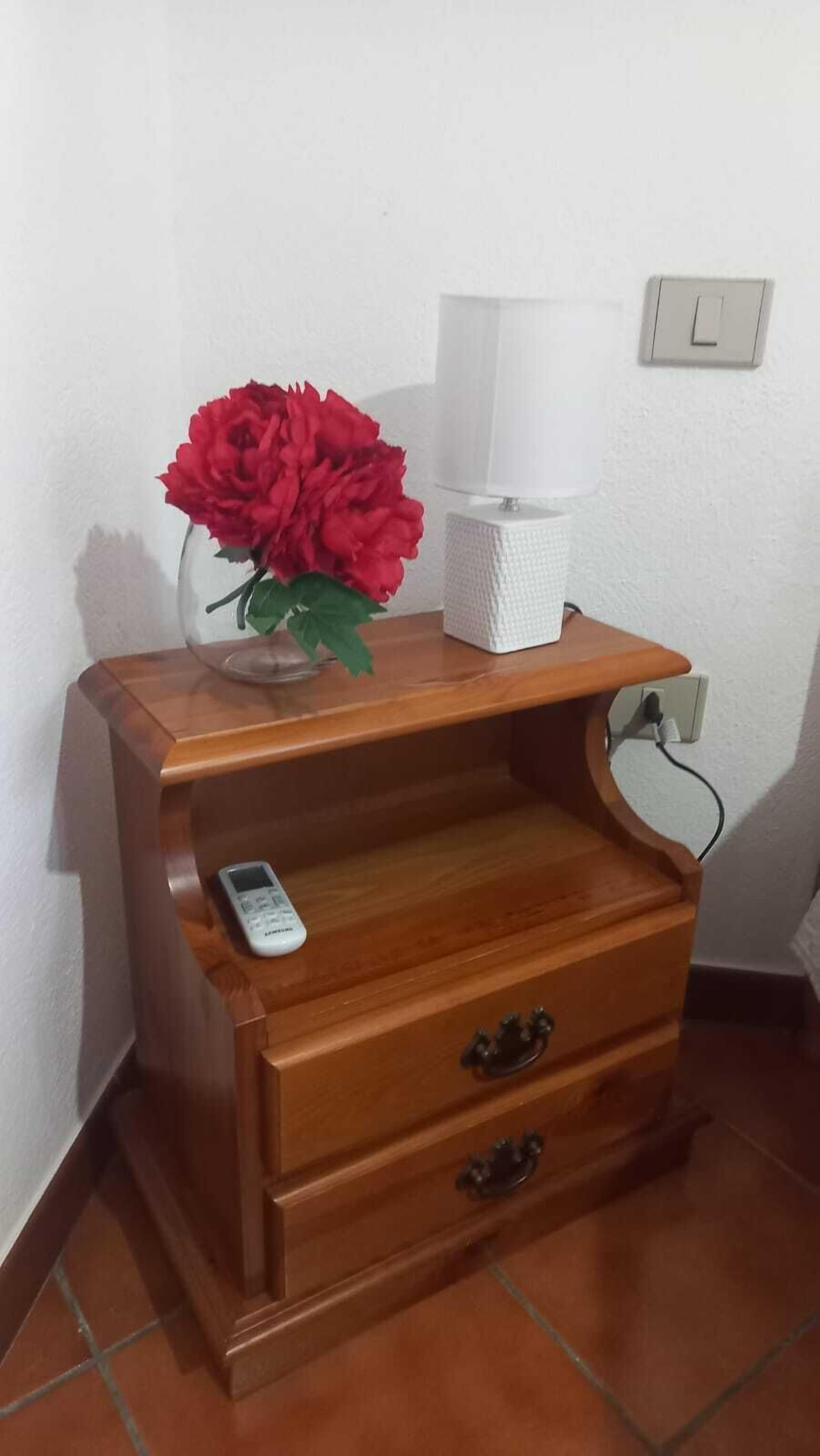 Una mesita de noche de madera con una lámpara, un control remoto y un florero con flores rojas.