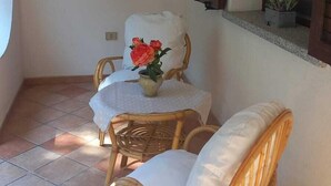 Una habitación con una silla de mimbre, una mesita con mantel blanco, una planta en maceta y una ventana con contraventanas.