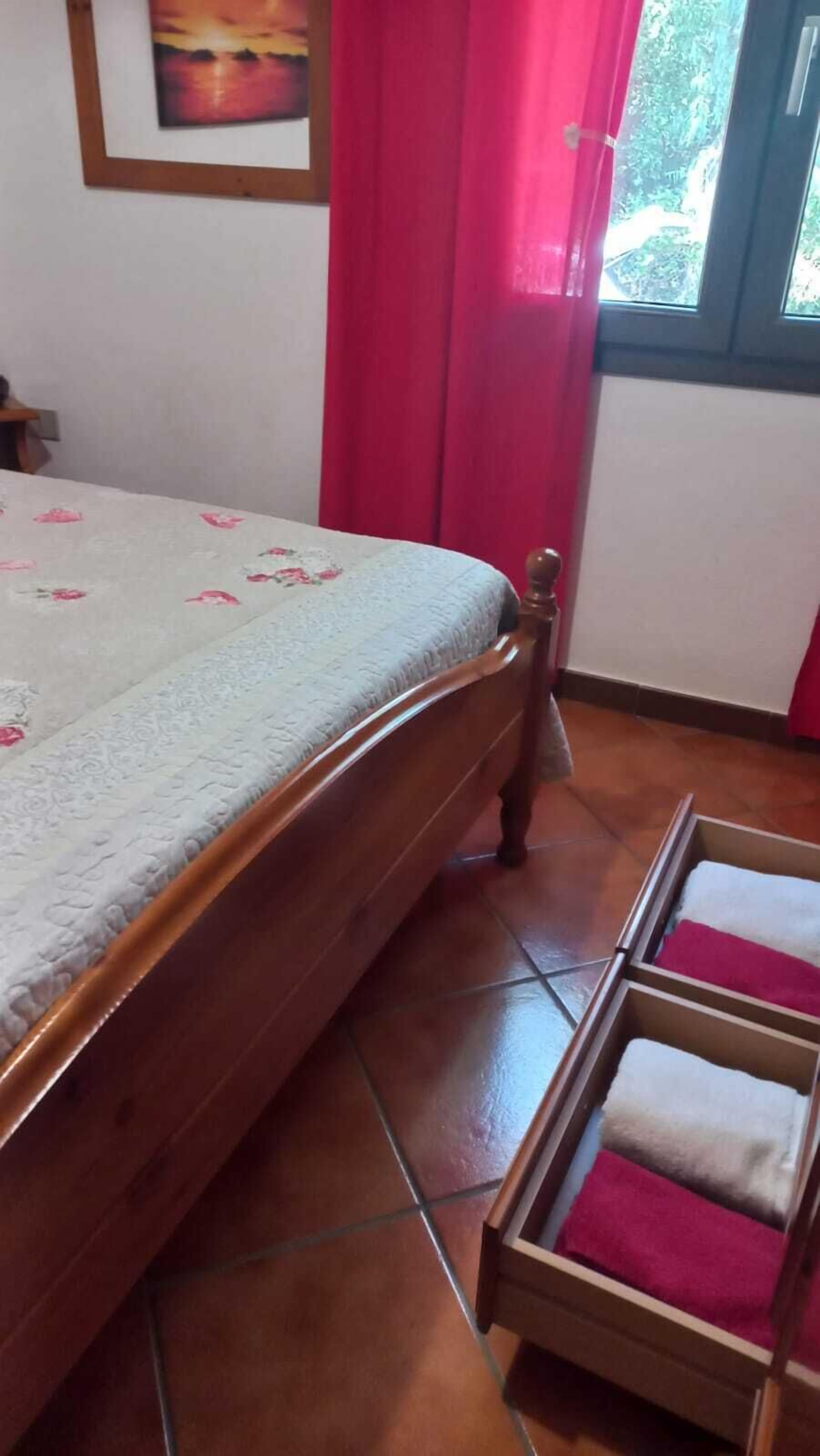 A bedroom with a bed, a wooden headboard, a window with curtains, and a tiled floor.