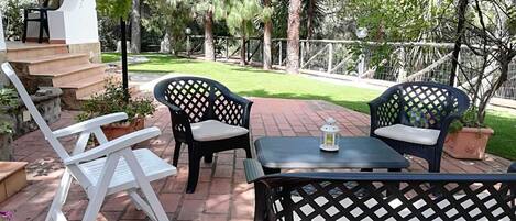 An outdoor patio area with a table and two chairs, surrounded by a brick pathway and greenery.