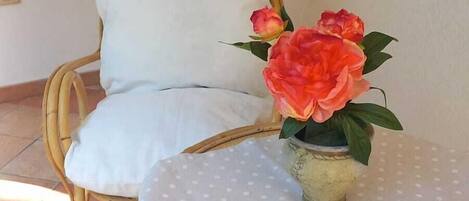 Una silla de mimbre con un cojín blanco, una mesita con mantel de lunares y un jarrón con flores.