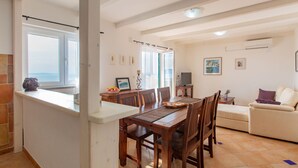Un espace repas comprenant une table et des chaises en bois, un canapé et un téléviseur. On y trouve des fenêtres avec des rideaux, une cheminée et un sol carrelé.