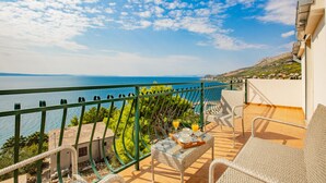 Un balcon agrémenté de meubles en osier, une table sur laquelle est disposée une sélection de produits pour le petit-déjeuner, et une vue panoramique sur la mer et les collines.