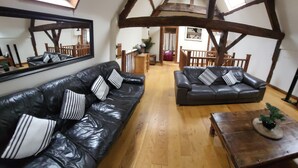 Ein geräumiges Wohnzimmer mit hohen Decken und freiliegenden Holzbalken. Es gibt zwei schwarze Ledersofas mit gestreiften Kissen, einen Holztisch und einen großen Spiegel an der Wand.