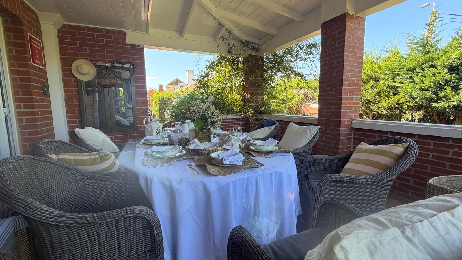 A covered patio with a table set for dining, wicker furniture, and a brick wall.