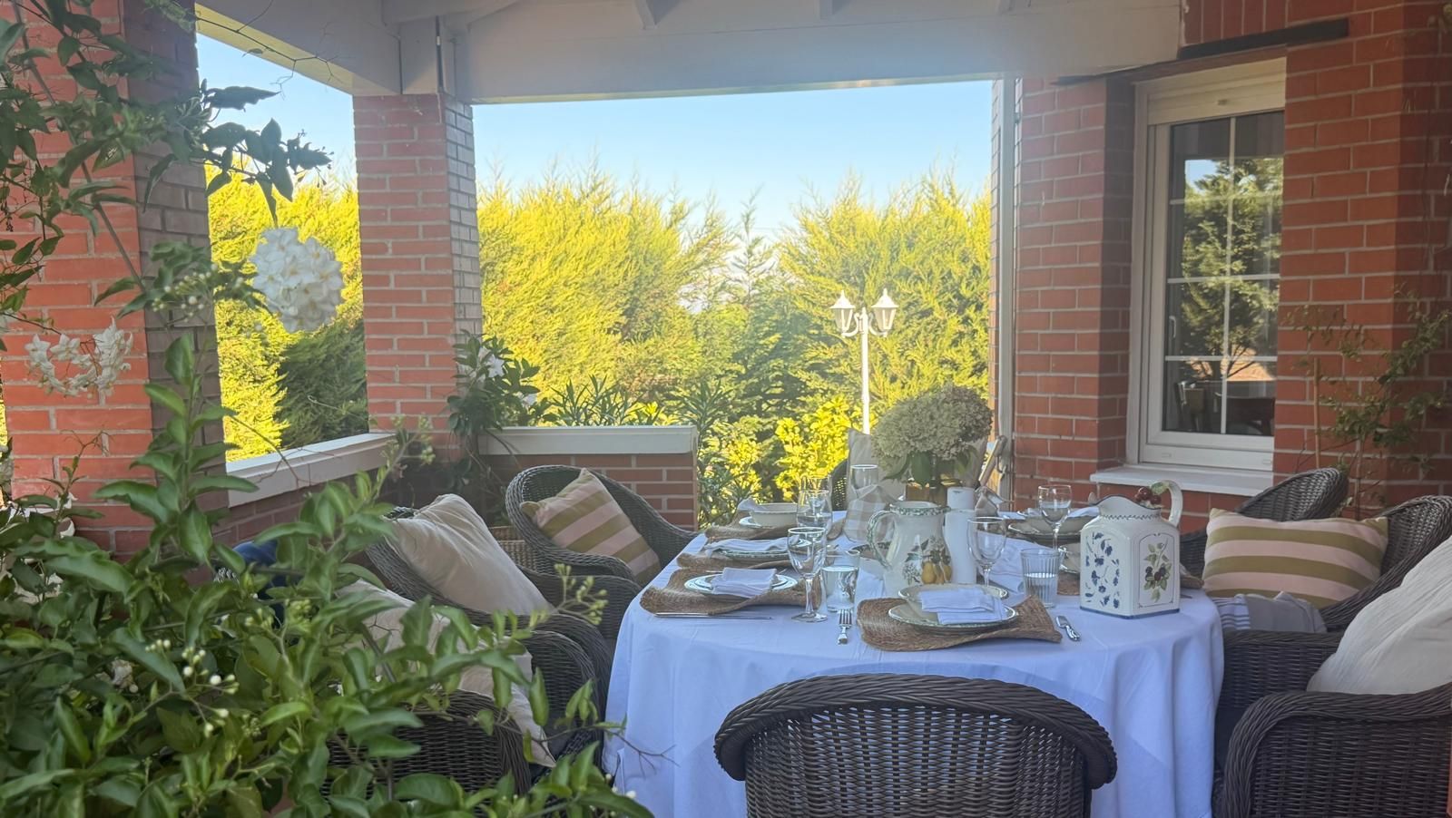 An outdoor dining area with wicker furniture and a table set for a meal, surrounded by brick walls and greenery.