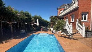 A swimming pool with a tiled deck, surrounded by lounge chairs and a brick house with a balcony.