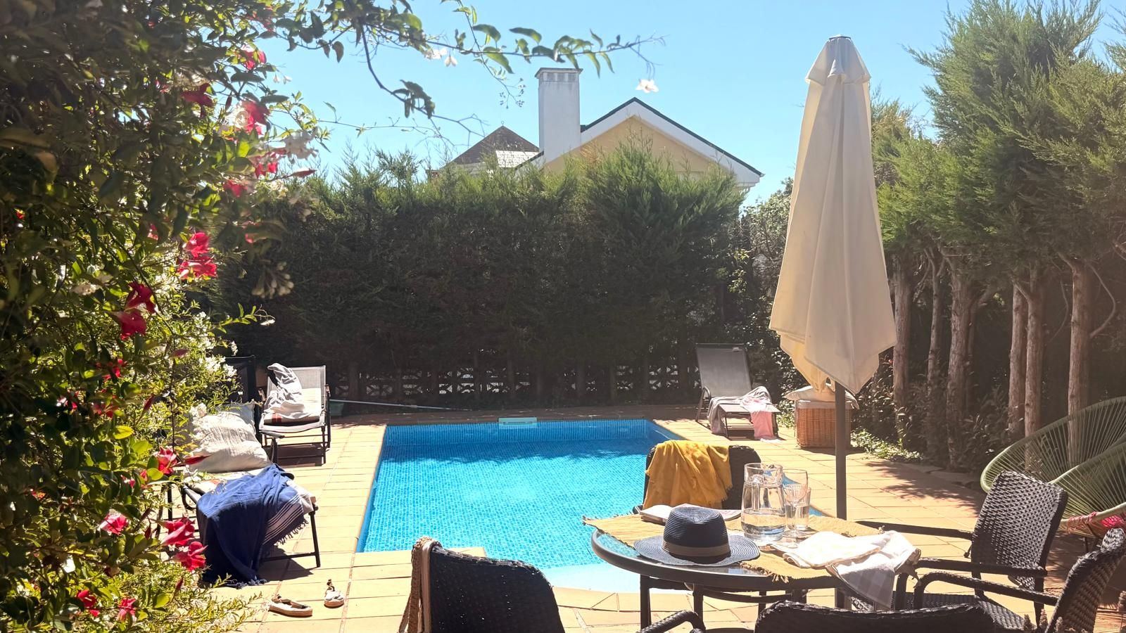 Ein Swimmingpool mit Liegestühlen und Sonnenschirmen, umgeben von viel Grün und einem Haus im Hintergrund.