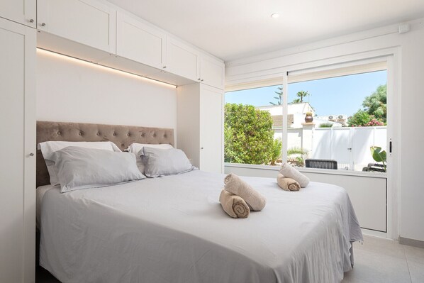 A bedroom with a large bed, white walls, and a view of greenery outside. - Villa Claudine,  800 meters from the beach with pool, for 6 People (Alcúdia)