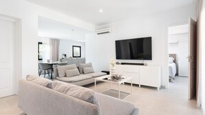 A modern living room with a grey sofa, a flat-screen TV mounted on the wall, and a minimalist kitchen in the background. - Villa Claudine,  800 meters from the beach with pool, for 6 People (Alcúdia)