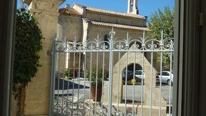 A gated entrance with a wrought iron gate, a stone pillar, and a building with a tiled roof and bell tower in the background. - Lovely house, garden with pool in tranquil village near coast and countryside (Raissac-D'aude)
