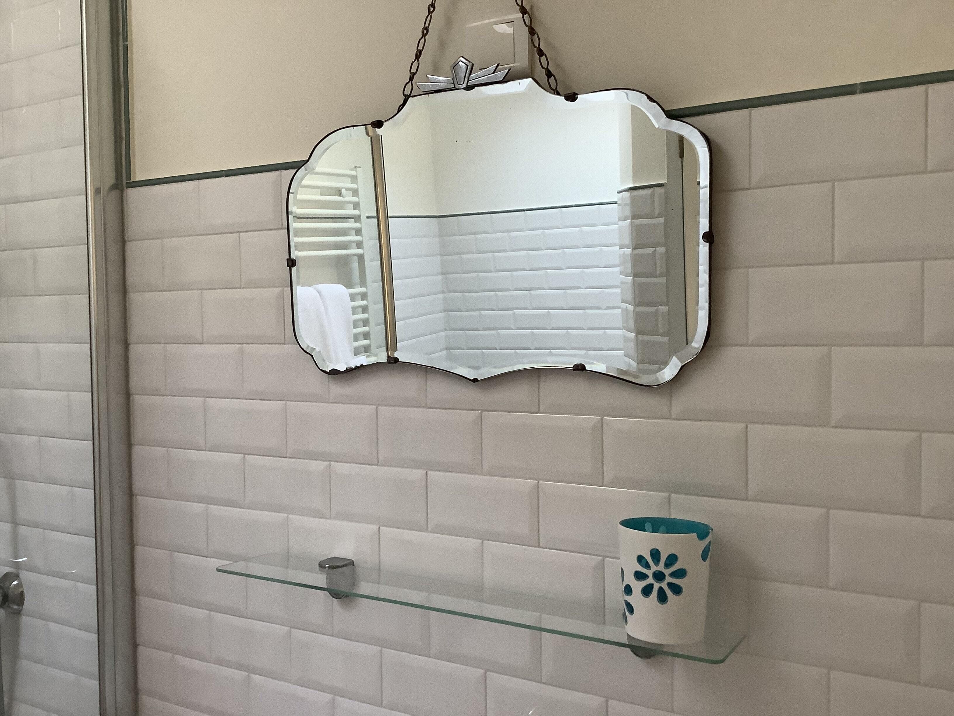 A bathroom with a white tiled wall, a glass shelf with a cup, and a mirror with a unique shape.