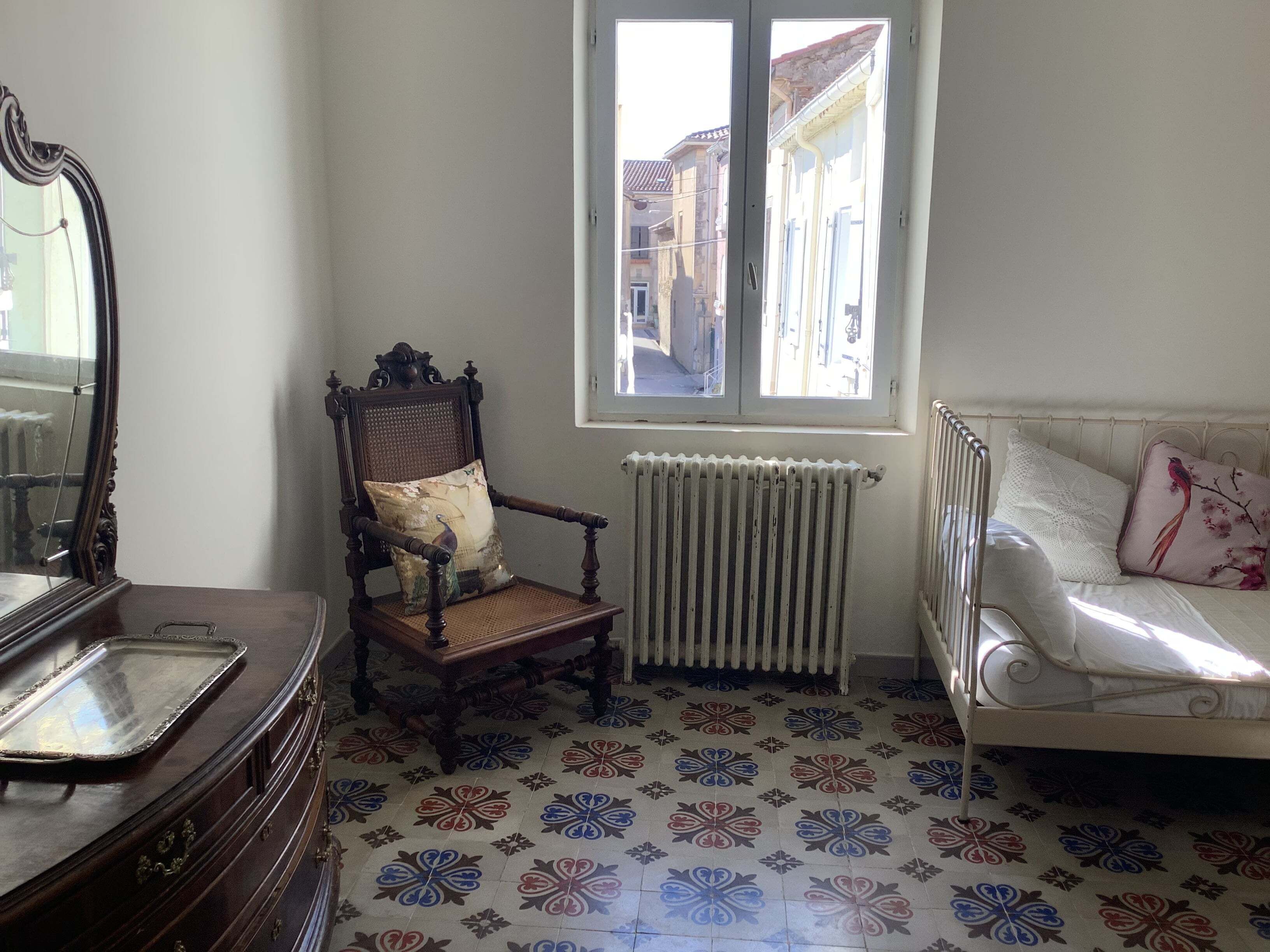 A room with a wooden chair, a radiator, a window with a view of buildings, and a bed with a pillow.