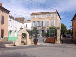 A street view with a gated entrance, a fountain, and a building with multiple windows and a tiled roof. - Lovely house, garden with pool in tranquil village near coast and countryside (Raissac-D'aude)