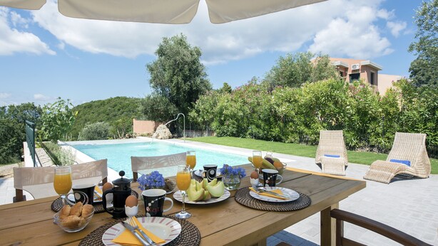 Mesa de desayuno dispuesta con platos, tazas y una cafetera, con vista a una piscina y abundante vegetación.