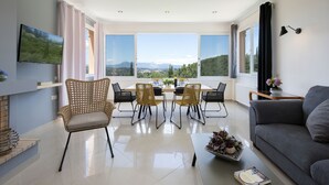 Una sala de estar moderna con comedor, ventanas grandes con vistas a la montaña, un televisor de pantalla plana y un sofá gris.