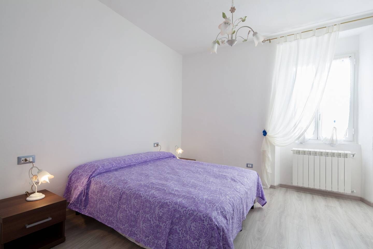 Ein Schlafzimmer mit Bett, Nachttischlampen, einem Heizkörper und einem Fenster mit Vorhängen.