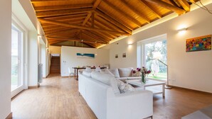 Een ruime woonkamer met een hoog houten plafond, een witte bank, een salontafel en uitzicht op het landschap door grote ramen.