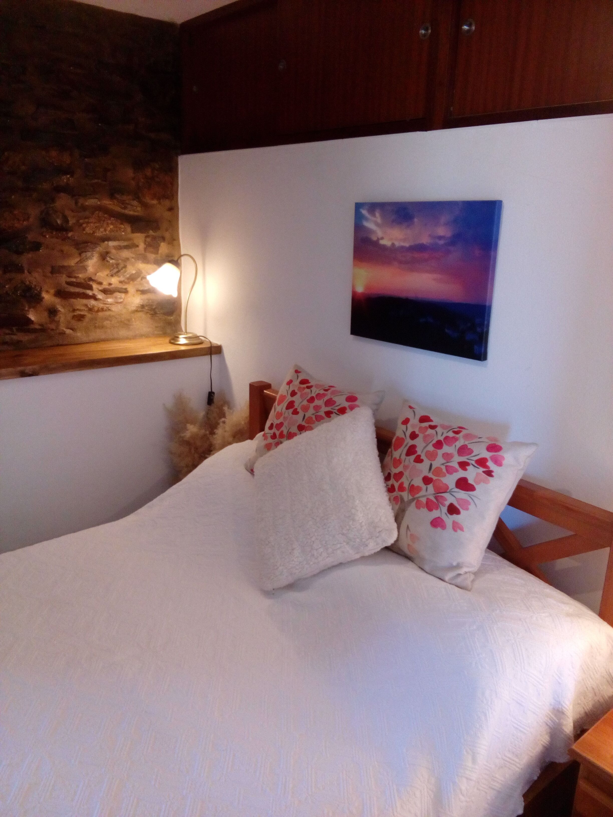 A bedroom with a stone wall, a painting of a sunset, and a bed with white bedding and heart-patterned pillows.