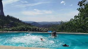Una piscina con una persona nadando, otra persona descansando con las piernas extendidas, y vistas del paisaje circundante.