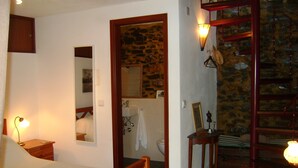 Un dormitorio con cama de madera, mesita de noche, lámpara de noche, baño con lavamanos, espejo y toallero.