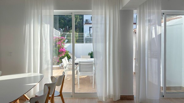 A modern interior with a dining area, white curtains, and a view of an outdoor patio with chairs and a table.