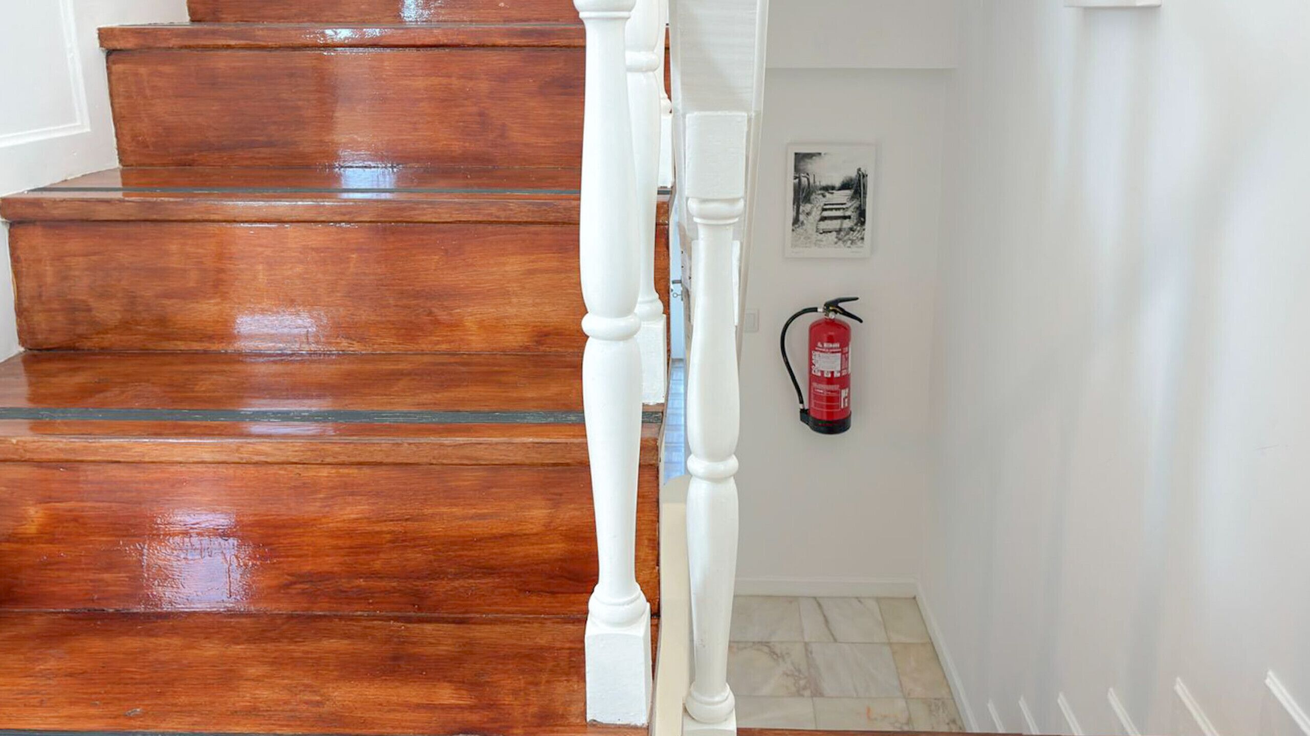 Un escalier en bois avec des balustrades blanches, un extincteur accroché au mur et un tableau encadré.