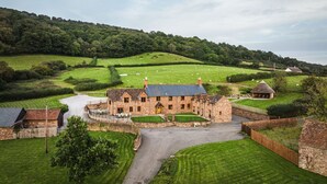 A stone building with a thatched-roof structure, surrounded by well-maintained lawns and a rural landscape. - Luxury Farmhouse with Stunning Somerset Views (Taunton)
