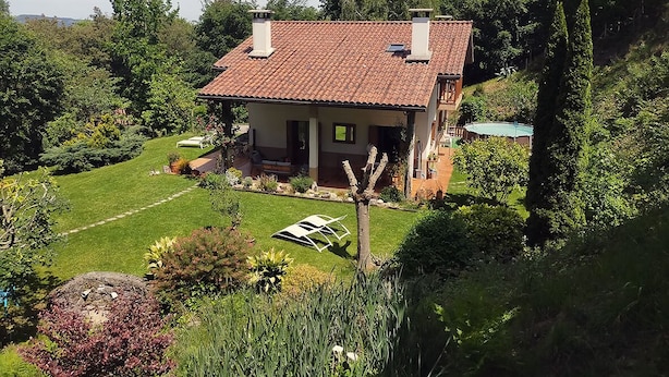 A two-story house with a red-tiled roof, surrounded by lush greenery and a well-maintained lawn.