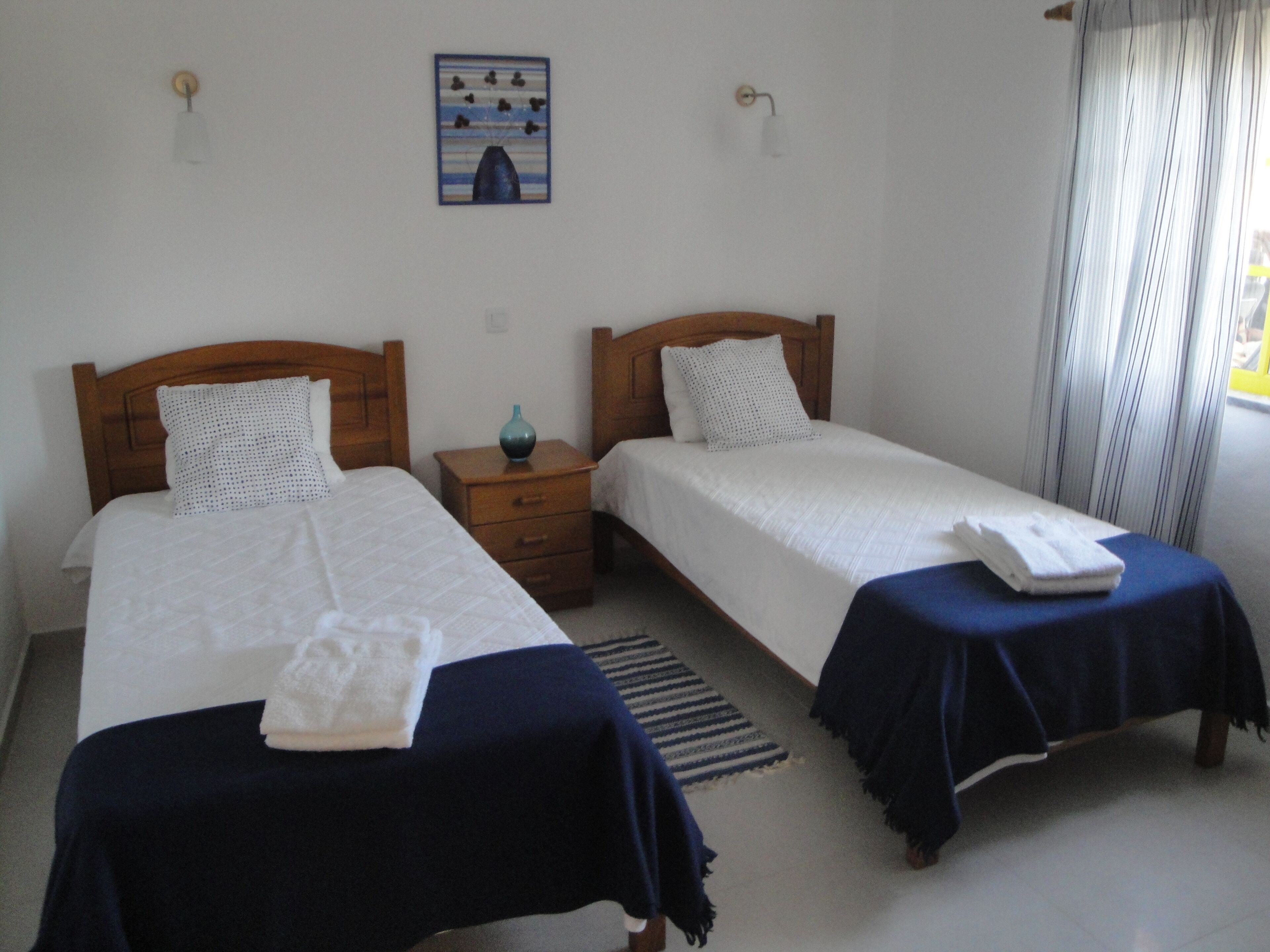 A hotel room with two single beds, a wooden nightstand, a small table, and a wall-mounted painting.
