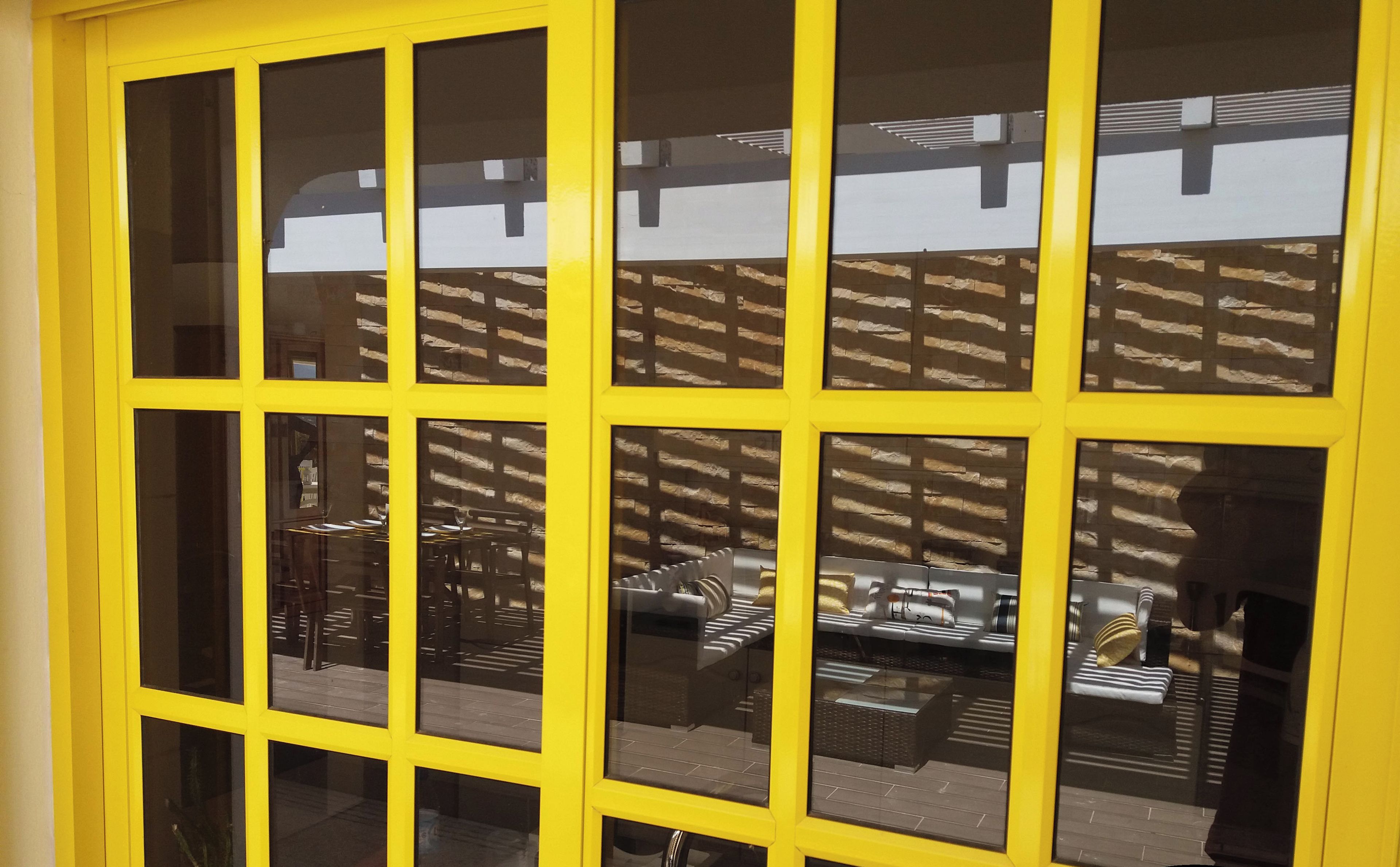 Ein gelb gerahmtes Fenster mit vielen Sprossen, das einen Innenraum mit Möbeln und einer Ziegelwand reflektiert.