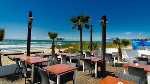 An outdoor dining area with wooden tables and chairs, overlooking a beach with palm trees and the ocean. - 75m to Beach Luxury 4-Bed Villa with Heated Pool & Oasis Patio in Marbella (Marbella)