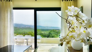 Una habitación con vistas a un paisaje verde a través de un ventanal grande, un florero blanco con flores blancas sobre una mesa, y un comedor con sillas y una mesa.