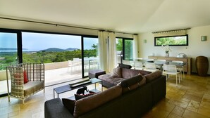 Una sala de estar moderna con un sofá grande, una mesa de comedor y sillas. Hay un balcón con vista al paisaje.