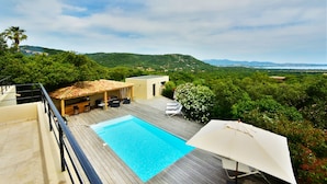 Una piscina en la azotea con terraza de madera, mobiliario exterior y vistas a la vegetación y colinas circundantes.