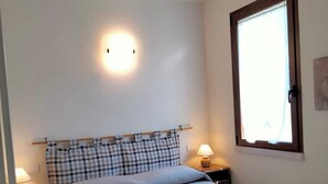 A neatly made bed with a checkered headboard, a bedside lamp, and a window with a wooden frame. - Villetta Gelsomino, in Bardolino Calmasino villa in residence with swimming pools. (Calmasino)