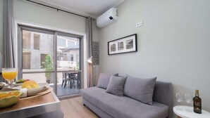 A modern living room with a grey sofa, a dining table with fruit and a glass of orange juice, and a small round table with two wine glasses and a bottle.