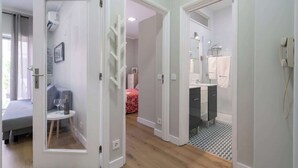 A hotel room with a bed, a small table with a bottle and glasses, a bathroom with a sink and mirror, and a hallway leading to another room.