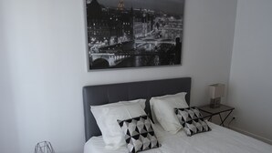 En snyggt bäddad säng med vita sängkläder och två mönstrade kuddar, ett litet runt bord med ett föremål i trådnät och en tavla med en stadsbild som hänger på väggen.