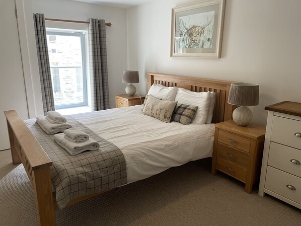 A neatly made bed with a checkered blanket, a bedside lamp, a wooden nightstand, and a drawer unit. - 2 bedroom first floor apartment at The Highland Club sleeping 6 via sofa bed. (Loch Ness)