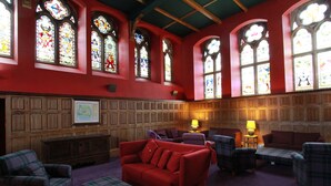 Una habitación con paredes rojas, paneles de madera y múltiples ventanas con vidrieras. Hay varios muebles, incluyendo un sofá rojo, sillones escoceses y una mesita con lámparas.
